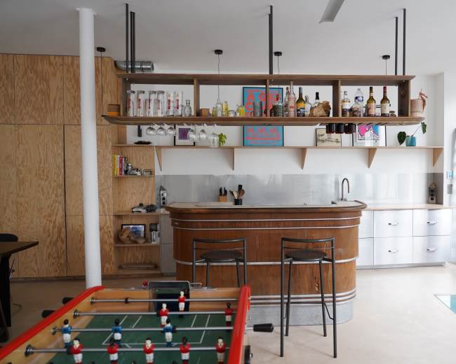 The scene features a modern kitchen and bar area with a wooden countertop, shelves displaying various bottles, and a foosball table in the foreground.