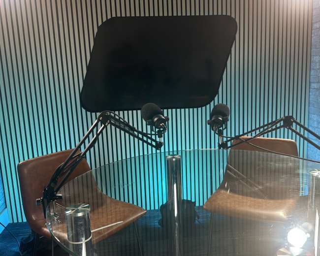 The image shows a modern podcast studio featuring two microphones mounted on arms, two brown chairs, and a glass table in front of a backdrop with vertical lines.