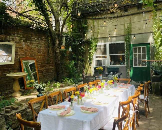 A long table is set for a meal in a garden with trees and string lights, surrounded by brick walls and green foliage.