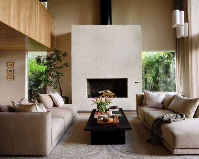 A spacious living room features neutral sofas, a central coffee table with flowers, and a large white brick fireplace, accented by expansive windows overlooking greenery.