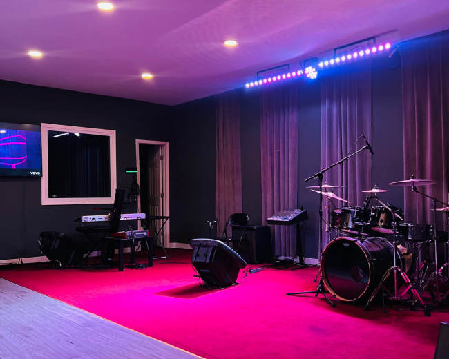 The image shows a music stage setup with a drum kit, keyboard, and amplifiers on a red carpeted floor, illuminated by purple and white stage lights.
