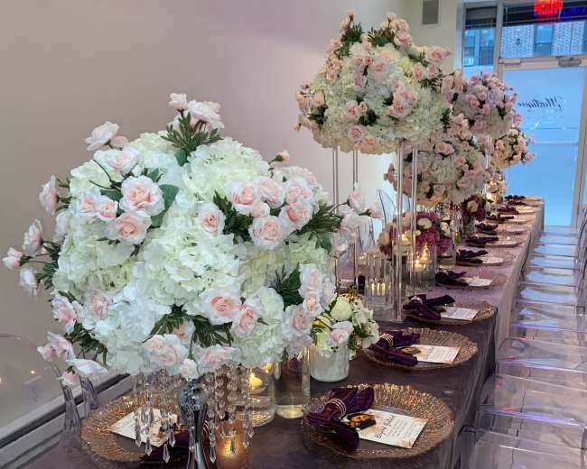 A long dining table is elegantly set with floral centerpieces, golden charger plates, and purple napkins.