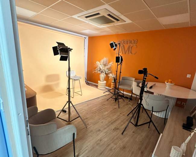 A studio setup features two camera stands with lights, a table with stools, and an orange accent wall.