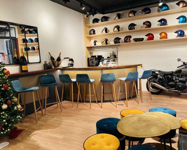 The image depicts a stylish bar area with blue stools, a motorcycle displayed on the right, and shelves lined with various motorcycle helmets.