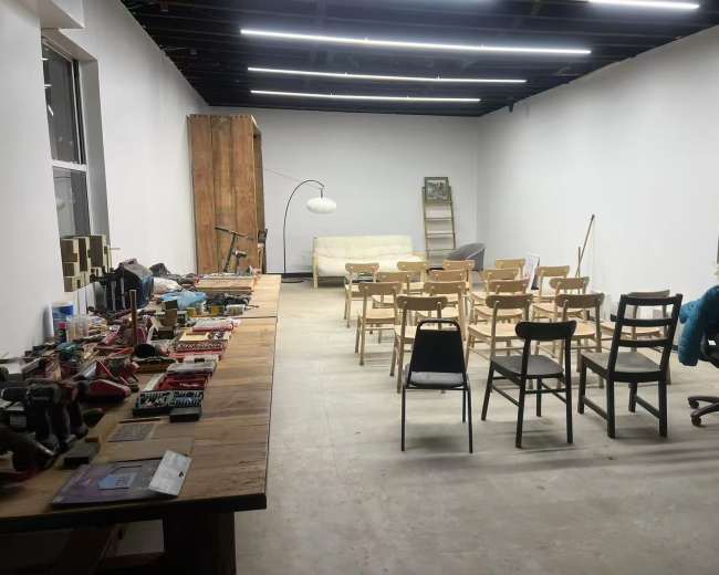 A spacious, unfinished room features a wooden table with various tools and supplies on one side and rows of wooden chairs facing a blank wall on the other.