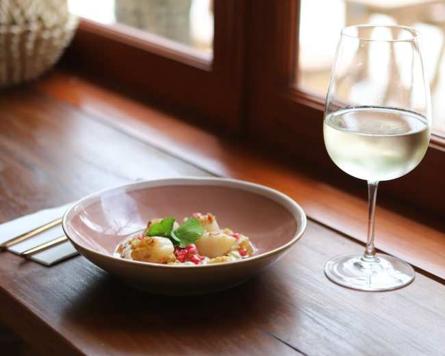A plate of food sits on a wooden table beside a glass of white wine, with large windows providing a view of the outdoors.