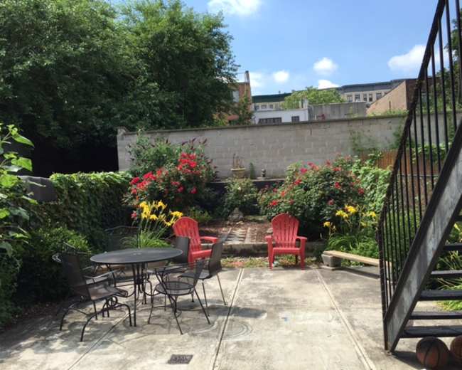 A garden space features a round black table and chairs surrounded by colorful flower beds and a stone pathway.