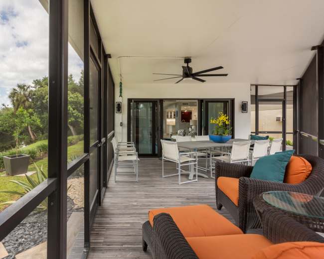 A screened-in porch features a dining table, comfortable seating, and a view of a green landscape with a body of water.