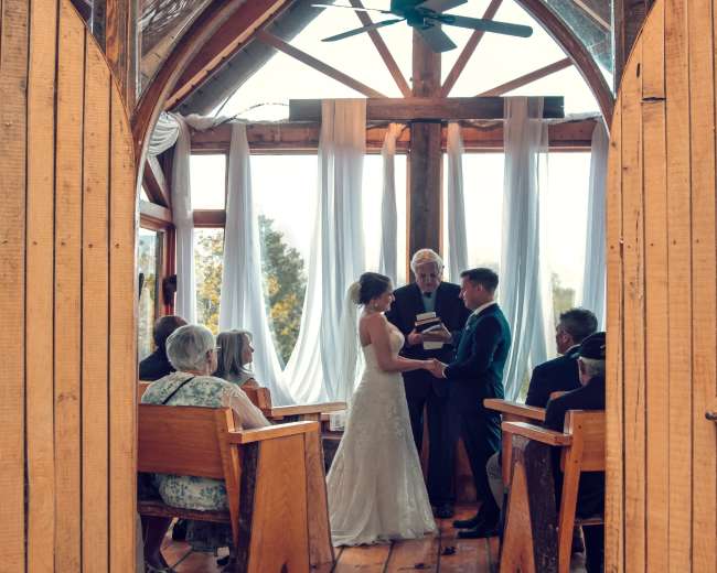 A couple stands at the altar inside a wooden chapel, surrounded by guests, as they exchange vows under a large ceiling fan.