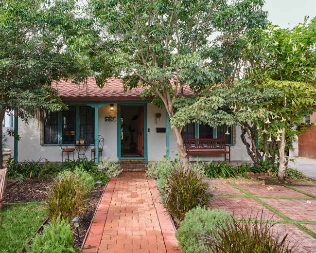 The image shows a single-story house with a red tiled roof, surrounded by green trees and landscaped gardens, with a brick walkway leading to the front door.