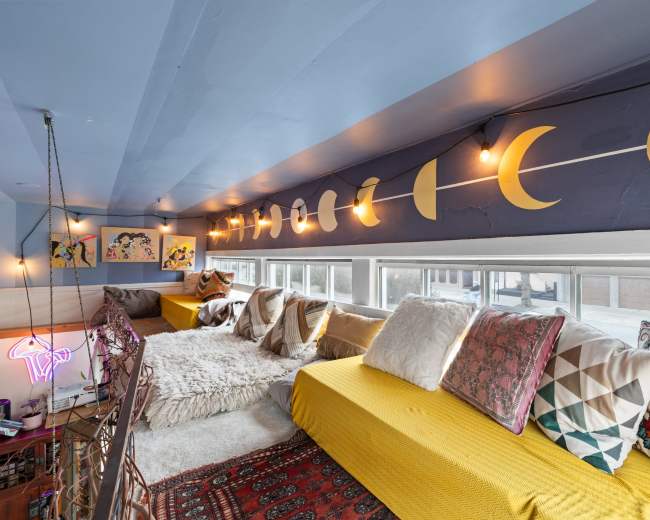 The image shows a cozy loft space with yellow seating adorned with various colorful pillows, illuminated by string lights and featuring moon-shaped decorations on the walls.