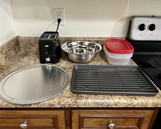 A kitchen countertop features a toaster, several metal bowls, a round baking pan, a cooling rack, and a plastic container with a red lid.