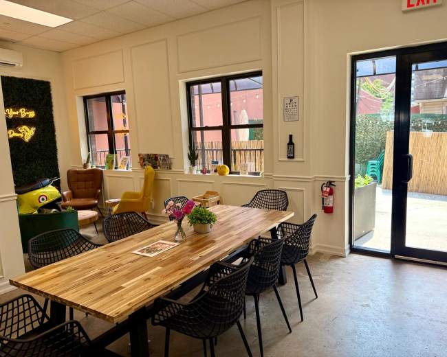 The image depicts a modern interior space with a wooden dining table surrounded by black chairs, large windows, and decorative elements like plants and seating areas.