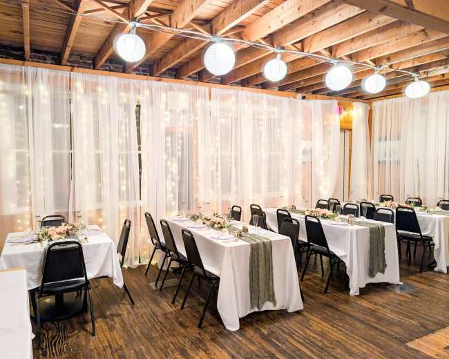 The image shows a warmly lit dining area with several tables covered in white linens and adorned with floral centerpieces, surrounded by sheer drapery and string lights.