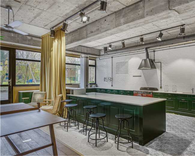 The image shows a modern kitchen with green cabinetry, a large island with barstools, and large windows providing natural light.