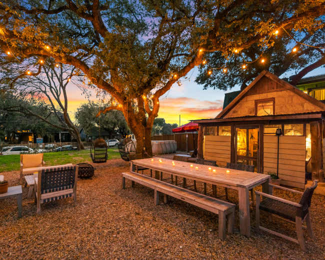 A rustic outdoor space features a long wooden dining table surrounded by benches, string lights illuminating the area, and a small shed in the background.