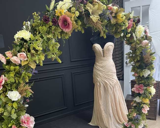 A floral arch surrounds a formal gown displayed on a mannequin against a dark wall.