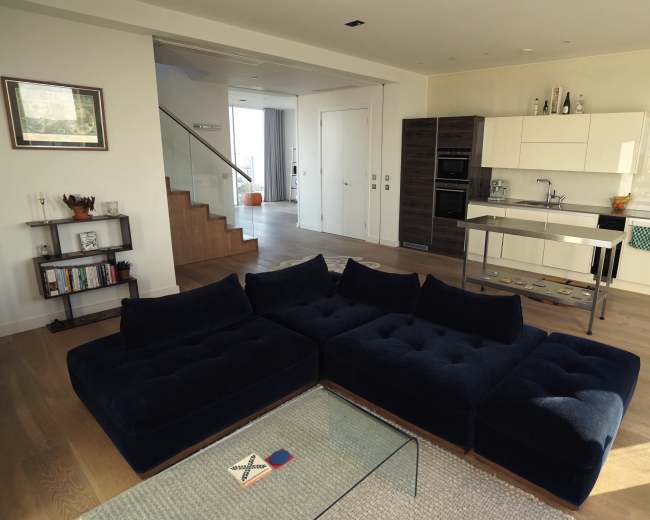 The image shows a modern open-concept living space featuring a large navy blue sectional sofa, a glass coffee table, and a kitchen area with white cabinetry and stainless steel appliances.