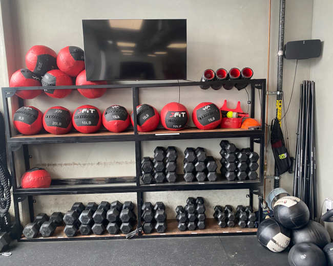 The image shows a well-organized gym storage rack containing various weights, including medicine balls and dumbbells, along with a television mounted on the wall.