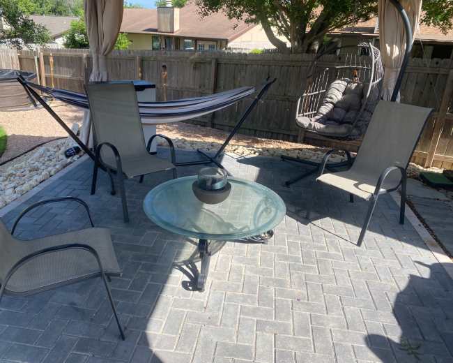 The image shows a patio area with a glass table surrounded by four chairs, a hammock in the background, and a hanging chair nearby.