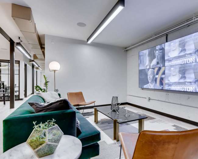 A modern lounge area features a large screen displaying video content, with a green sofa, two brown chairs, a glass table, and a floor lamp.