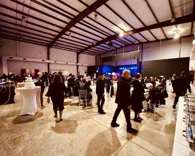 A large indoor venue hosts a gathering with guests mingling, tables set up, and a stage in the background.