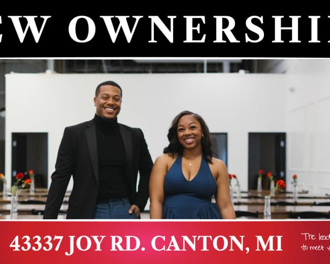 A man and a woman stand together in a modern event space, with tables decorated with roses and a banner announcing new ownership.