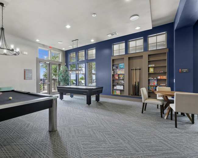 The image shows a recreational room with two pool tables, a dining area, and shelves filled with games, all set against a bright blue wall.