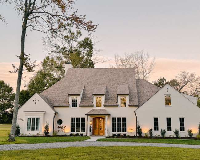 A large, modern white house with a steeply pitched roof and multiple windows is set in a landscaped yard surrounded by trees.