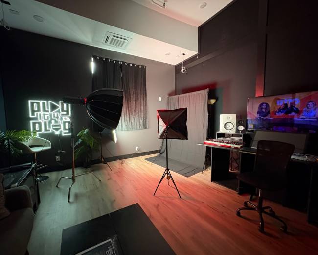 The image shows a modern media production studio with softbox lighting, a computer setup, and a neon sign on the wall.