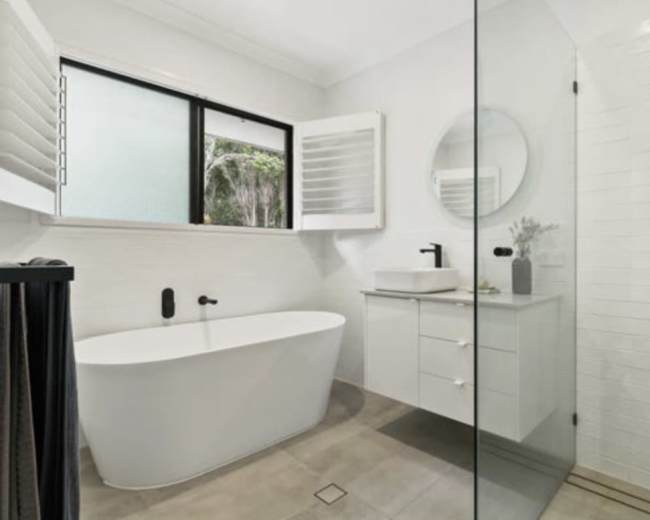 The image shows a modern bathroom featuring a freestanding bathtub, a glass shower enclosure, a wall-mounted sink with a mirror, and windows with louvered shutters.