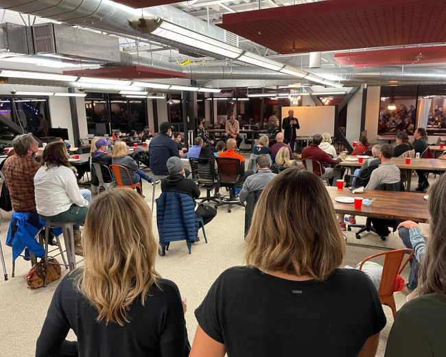 A group of people is seated in a large, open room, facing a speaker at the front, with tables and chairs arranged for an event.