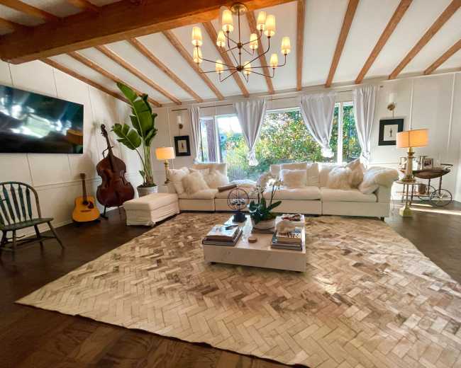 The living room features a large sectional sofa, a coffee table on a patterned rug, and a mix of decorative elements including a guitar, plant, and artwork, all under a chandelier with wooden beams overhead.