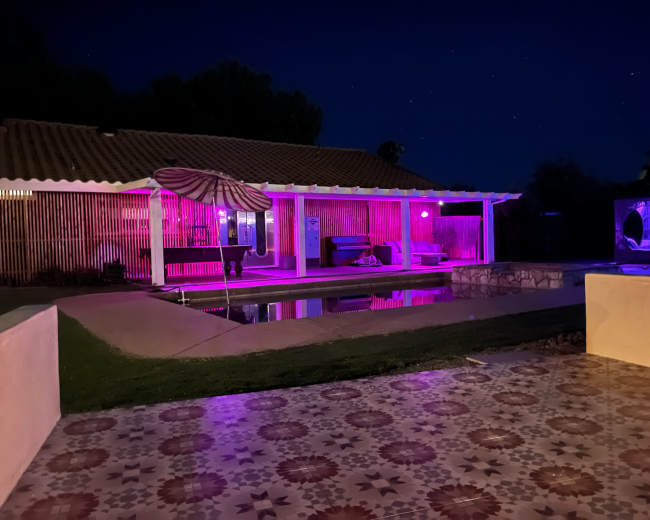 A modern outdoor area features a pool and patio illuminated by purple lights, with a house in the background.
