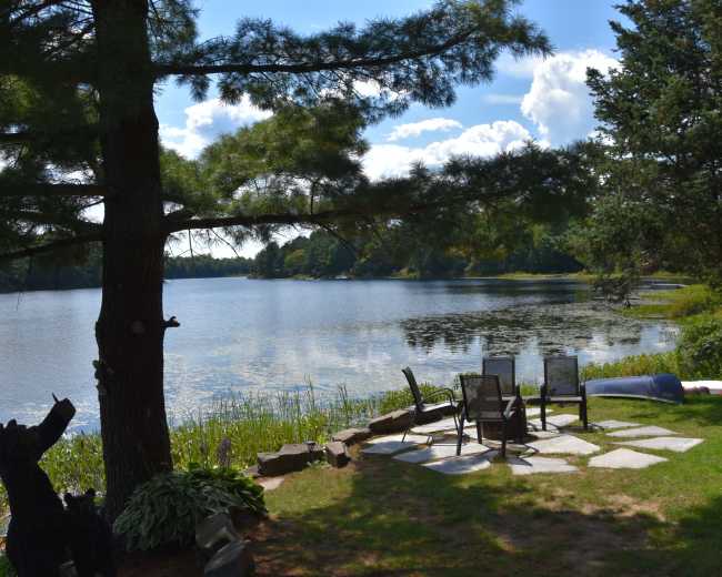A patio area with stone seating and table overlooks a calm lake surrounded by trees and greenery.
