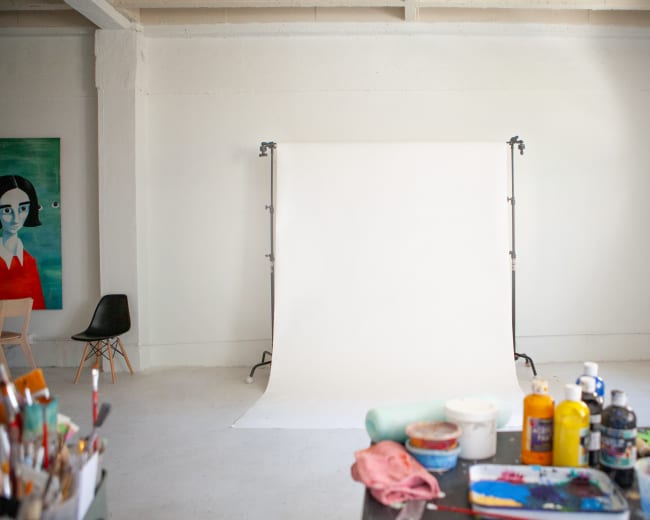 A well-lit studio space with a blank white backdrop, a chair, and art supplies scattered on a table.