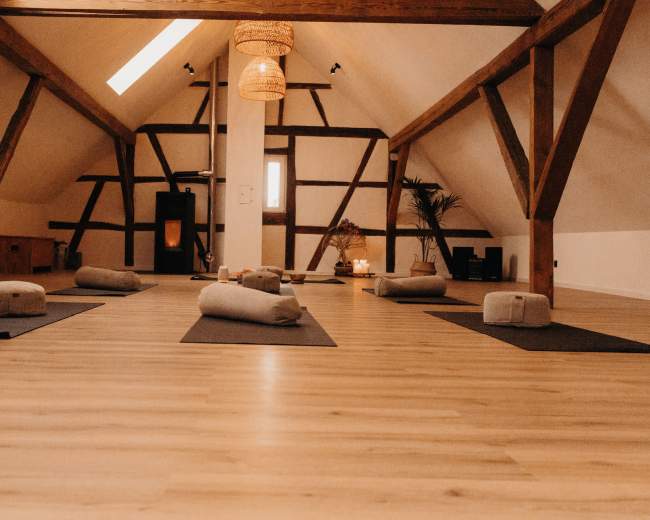 The image shows a spacious yoga studio with wooden beams, mats arranged on the floor, and meditation cushions set up for practice.