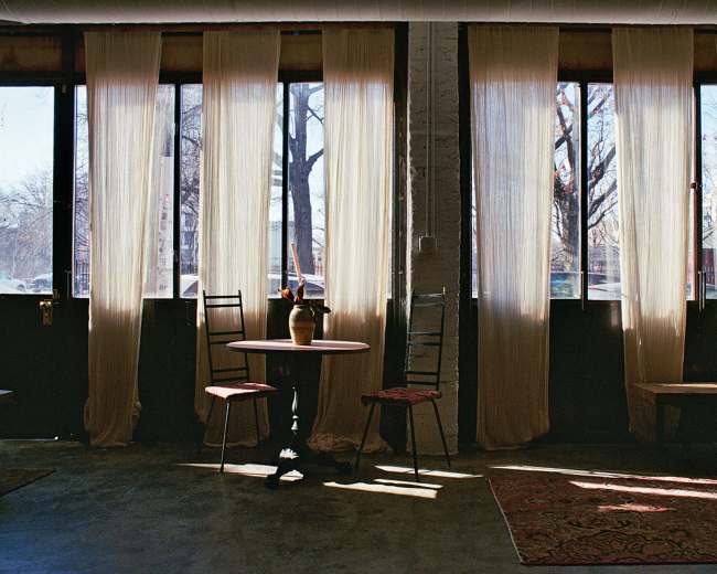 The image shows a room with large windows covered by sheer curtains, a small round table with a vase, and chairs arranged around it.