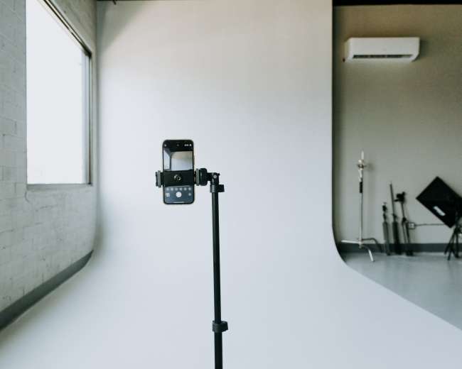 A smartphone is mounted on a stand facing a plain white backdrop in a photography studio.