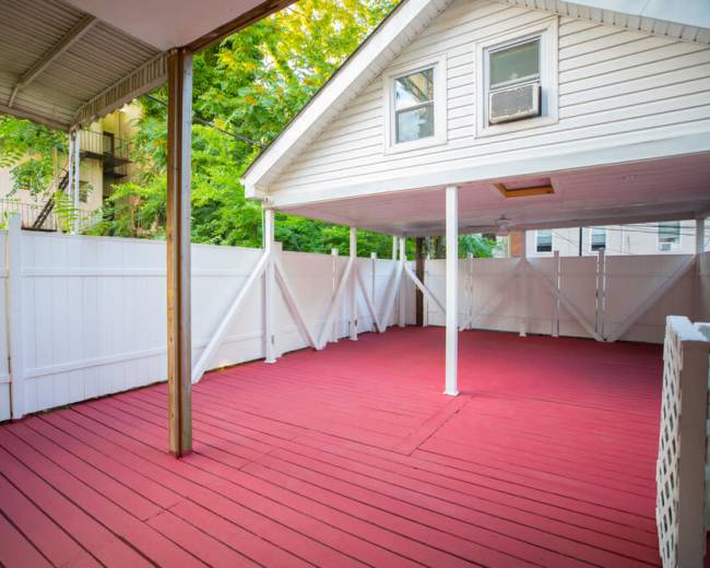 A covered outdoor space with a red wooden floor and white privacy fencing, surrounded by greenery.