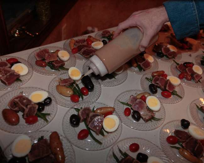 A person is squeezing a sauce from a bottle onto plates of food that feature sliced meat, olives, cherry tomatoes, and hard-boiled eggs.
