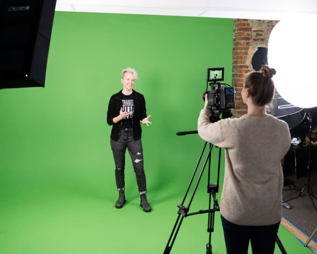 A person stands in front of a green screen while another person records them with a camera on a tripod.