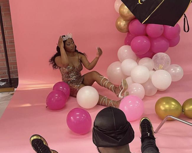 A model wearing a sparkly dress sits among pink, white, and gold balloons in front of a pink backdrop during a photoshoot.