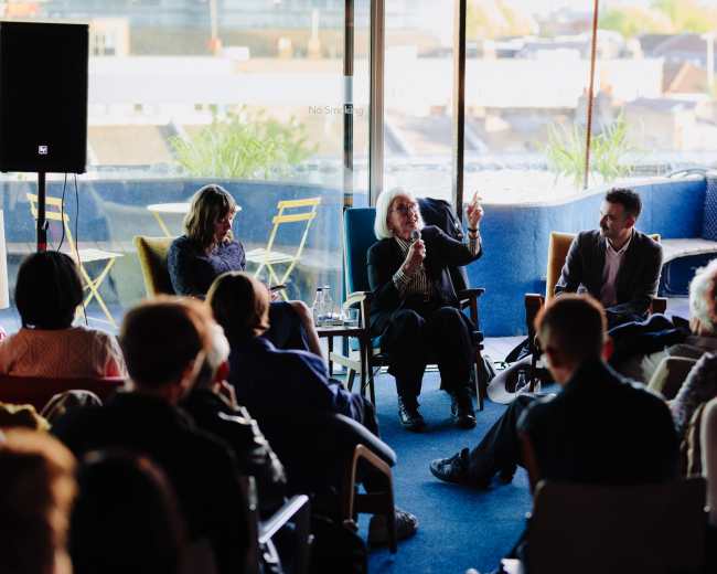 A panel discussion takes place in a well-lit room with large windows, featuring three speakers seated in chairs while an audience attentively listens.
