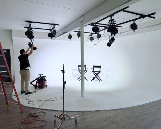 A person adjusts lighting equipment in a photography studio featuring a seamless white backdrop and two empty director's chairs.