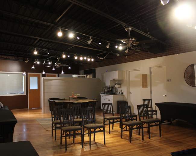 The image shows an indoor space with wooden floors, several chairs arranged in rows, a kitchenette area, and soft lighting from ceiling fixtures.