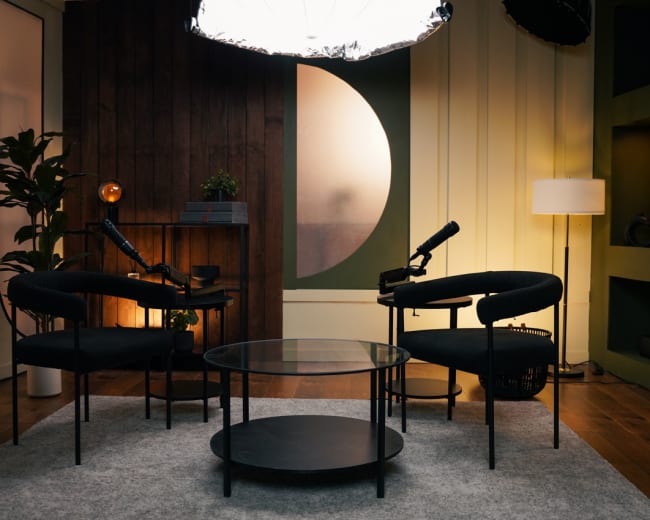 The image shows a cozy, modern interior with two black chairs facing a small glass table, under a large, circular light fixture.