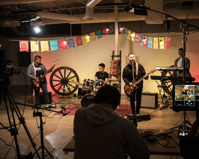 A band performs on a small stage with instruments while being filmed in a spacious room decorated with colorful banners.