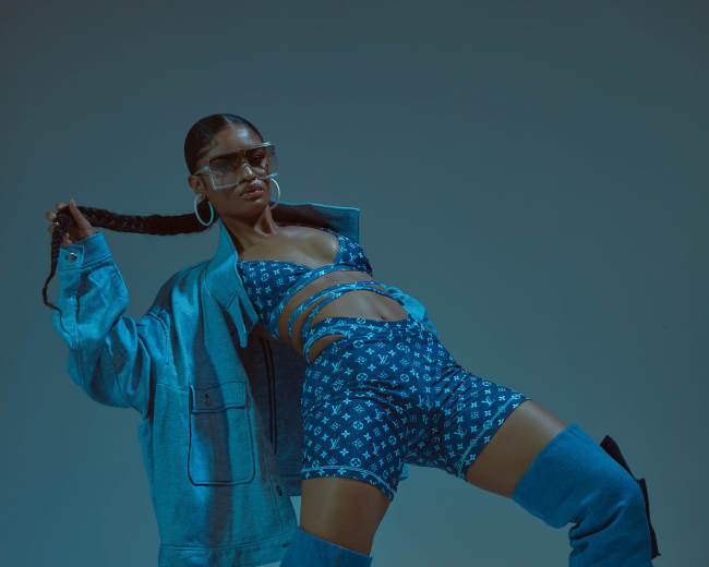 A model poses dynamically in a blue ensemble featuring a cropped top and shorts, complemented by oversized boots and a denim jacket.