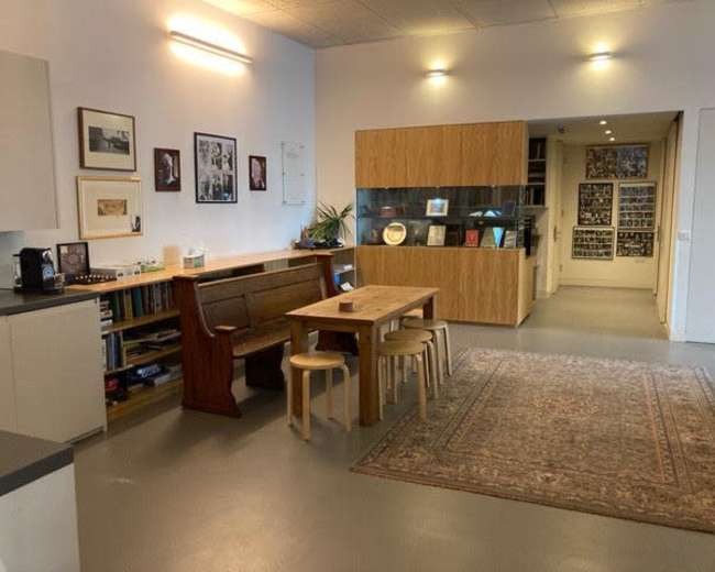 The image shows a spacious interior with a wooden dining table surrounded by stools, a modern cabinetry unit, and framed photographs on the walls.